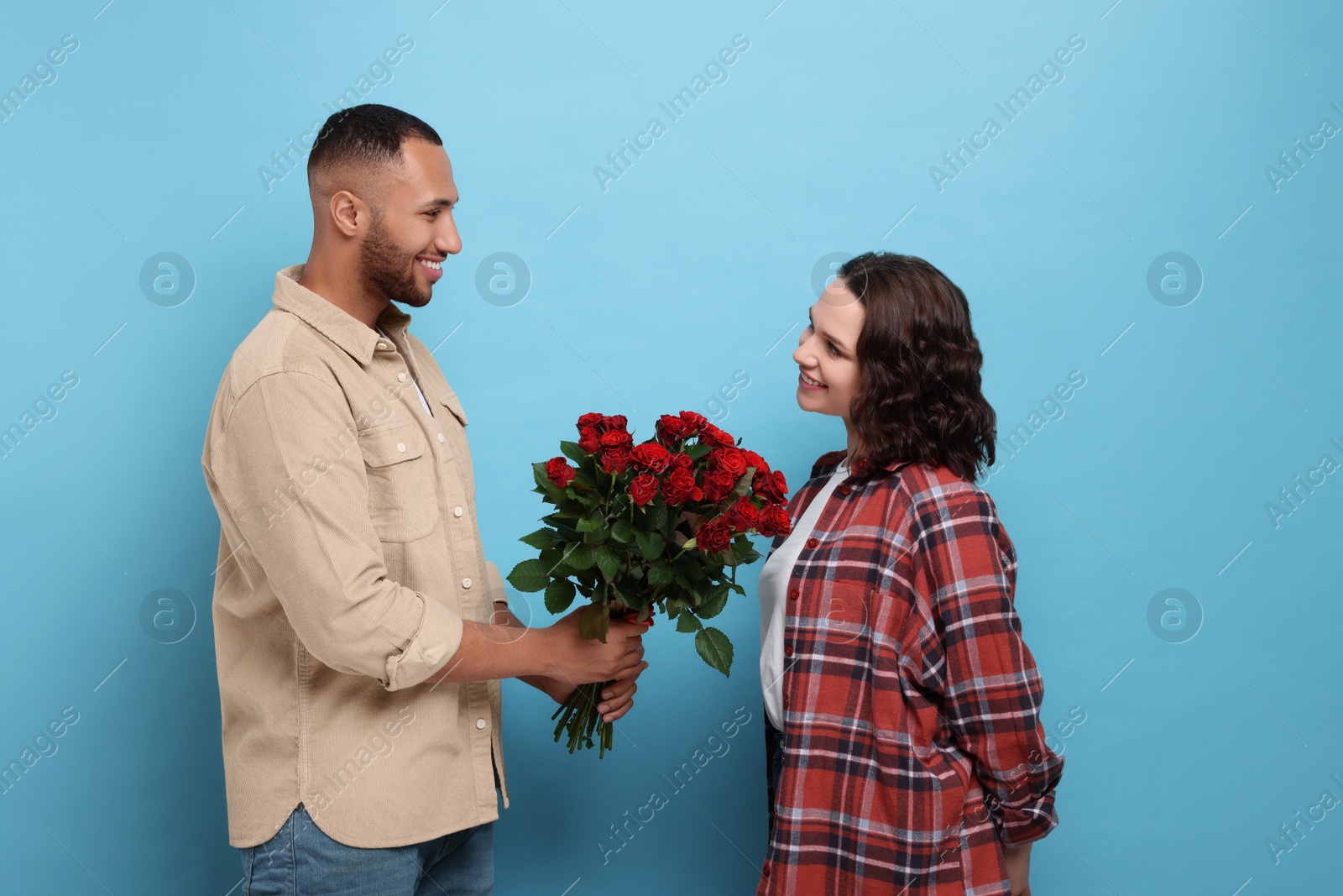 Photo of Boyfriend presenting bouquet of red roses to his girlfriend on light blue background. Valentine's day celebration