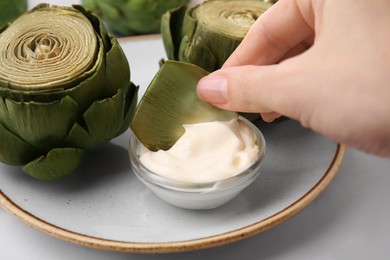 Woman dipping delicious cooked artichoke into sauce at white table, closeup