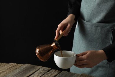 Turkish coffee. Woman pouring brewed beverage from cezve into cup at wooden table against black background, closeup. Space for text