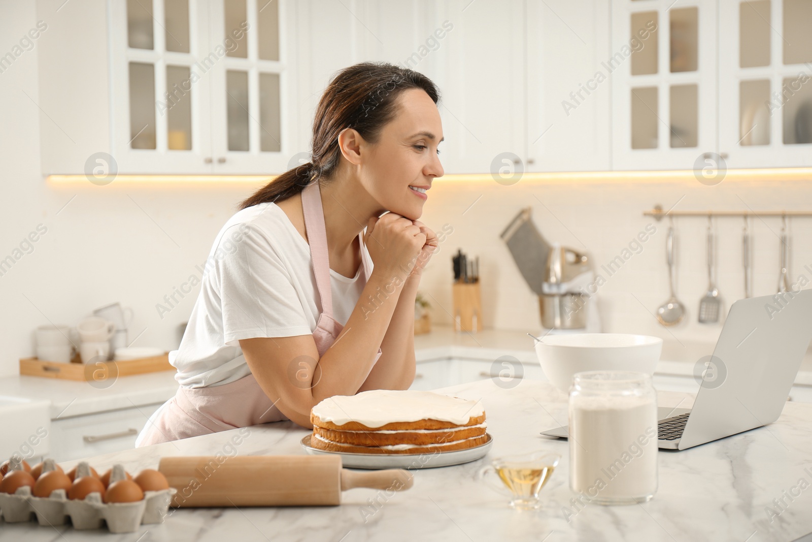 Photo of Woman making cake while watching online cooking course via laptop in kitchen