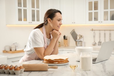 Photo of Woman making cake while watching online cooking course via laptop in kitchen