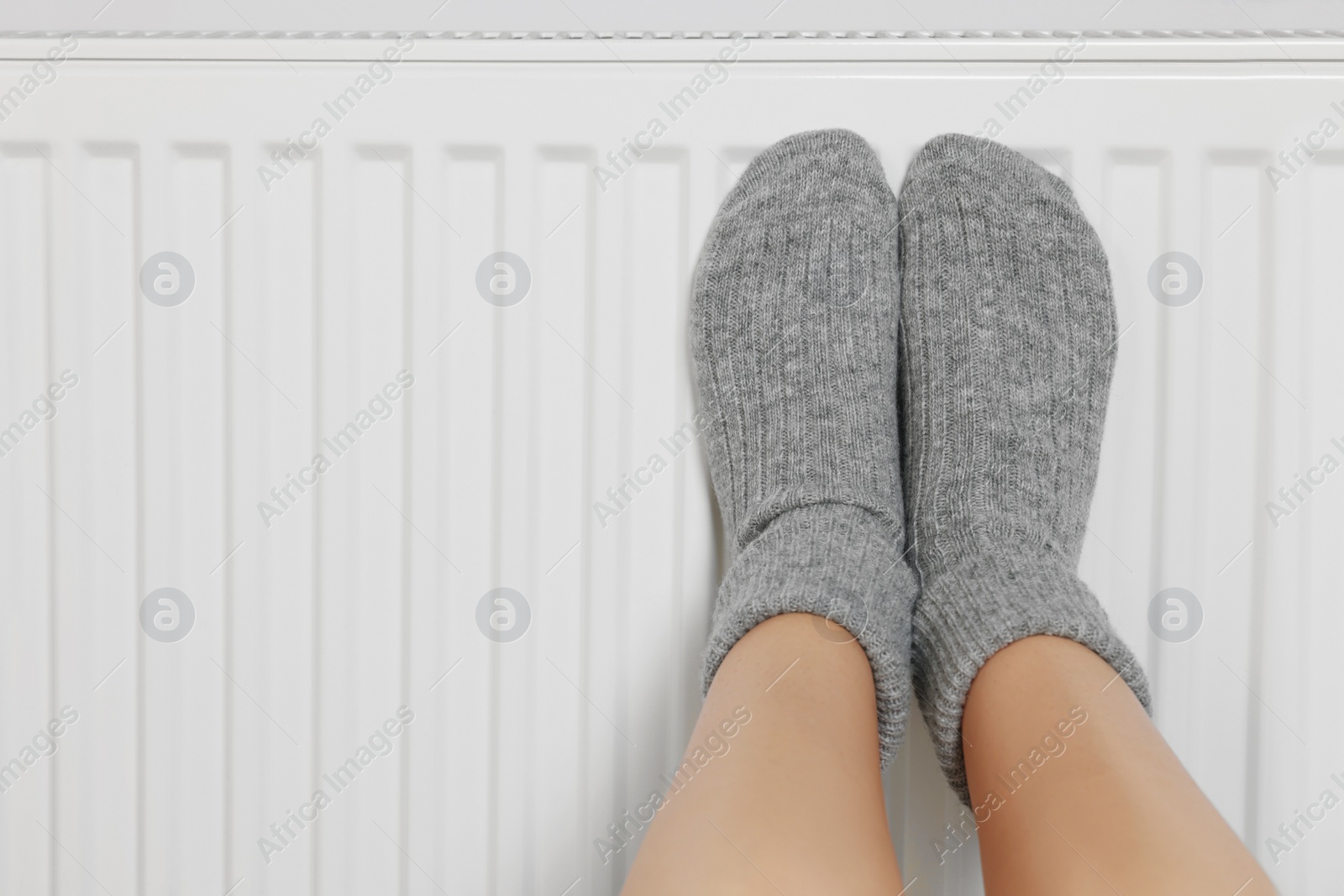 Photo of Woman warming legs on heating radiator near white wall, closeup. Space for text