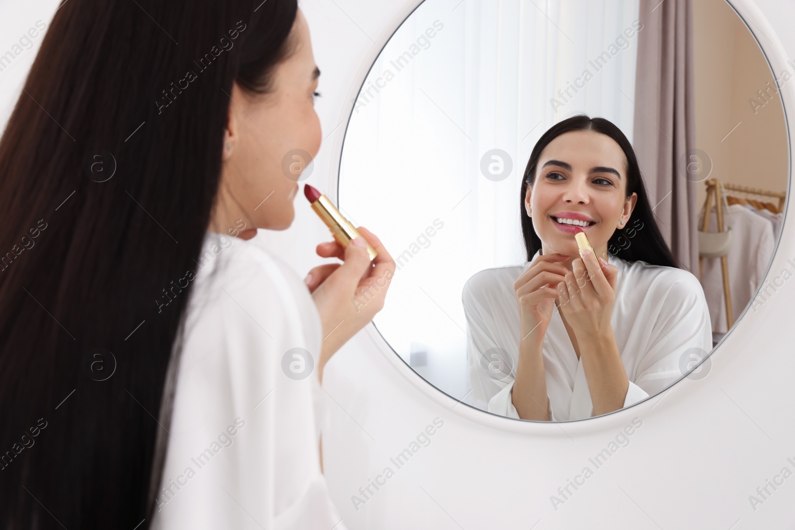 Photo of Beautiful young woman applying lipstick near mirror indoors