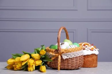 Easter basket with many painted eggs near tasty cake and bouquet of tulips on grey textured table. Space for text