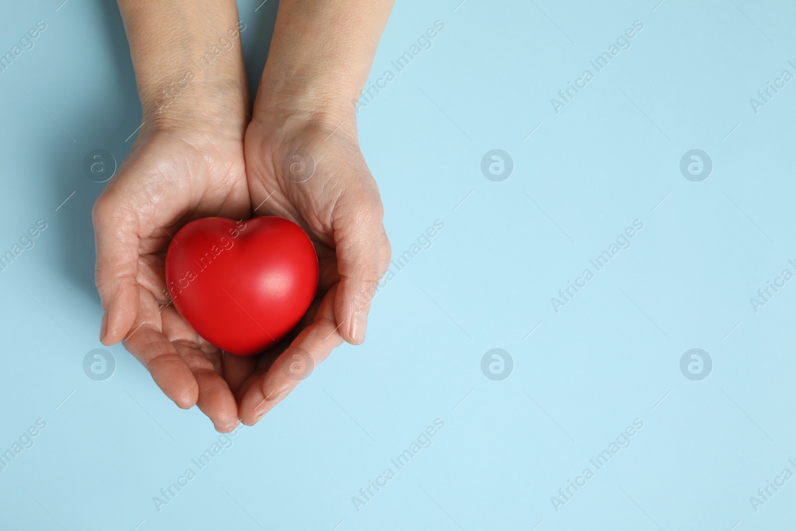 Photo of Elderly woman holding red heart in hands on light blue background, top view. Space for text