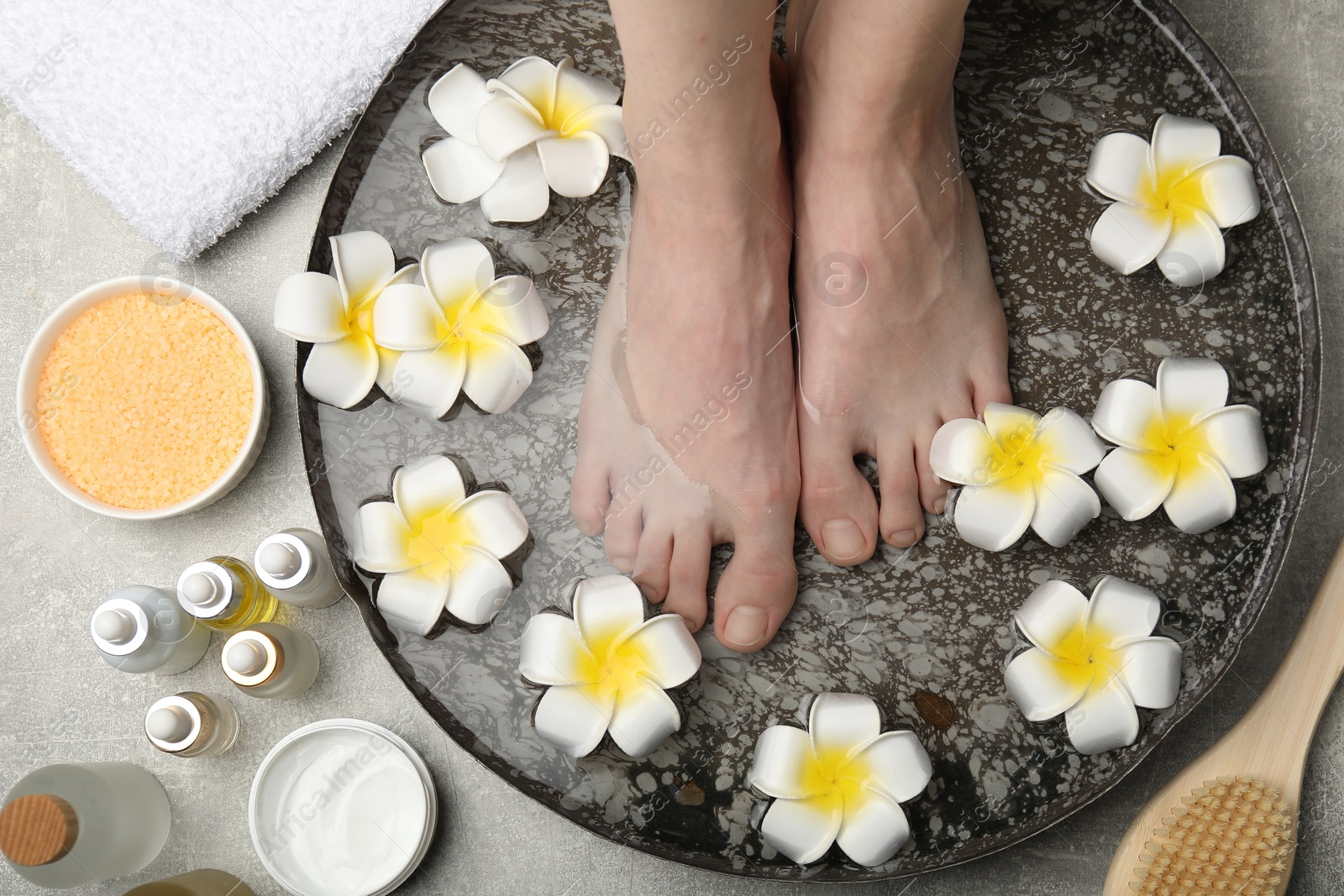 Photo of Woman soaking her feet in bowl with water and flowers on light grey floor, top view. Spa treatment