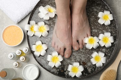 Photo of Woman soaking her feet in bowl with water and flowers on light grey floor, top view. Spa treatment