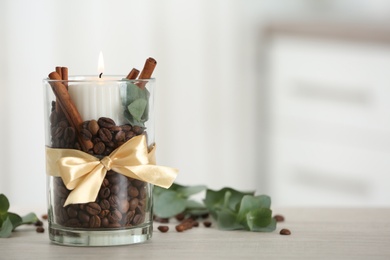 Photo of Burning candle in glass holder with coffee beans and cinnamon sticks on table indoors, space for text