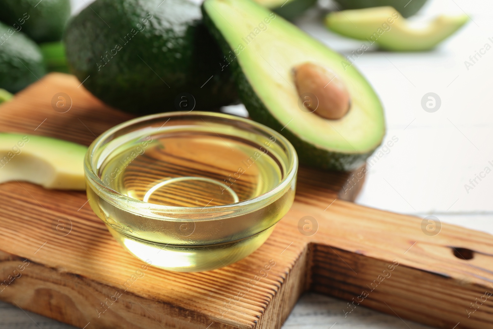 Photo of Board with bowl of natural oil and avocados on white wooden background, closeup. Space for text