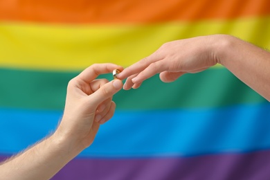 Photo of Young man putting wedding ring on his boyfriend's finger against rainbow background. Gay marriage