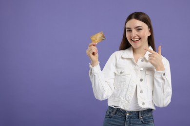 Happy woman pointing at credit card on purple background, space for text