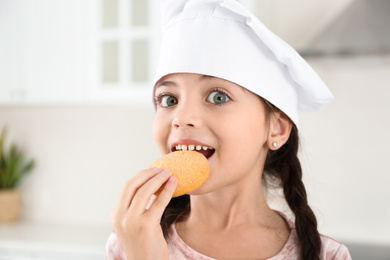 Photo of Cute little girl wearing chef hat eating cookies in kitchen