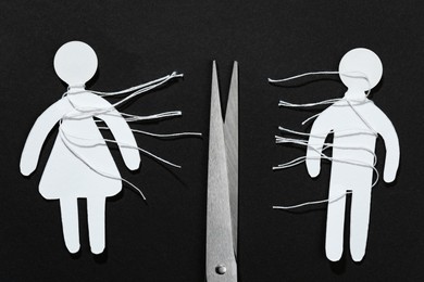 Photo of Divorce concept. Paper human figures with cut white threads and scissors on black background, flat lay