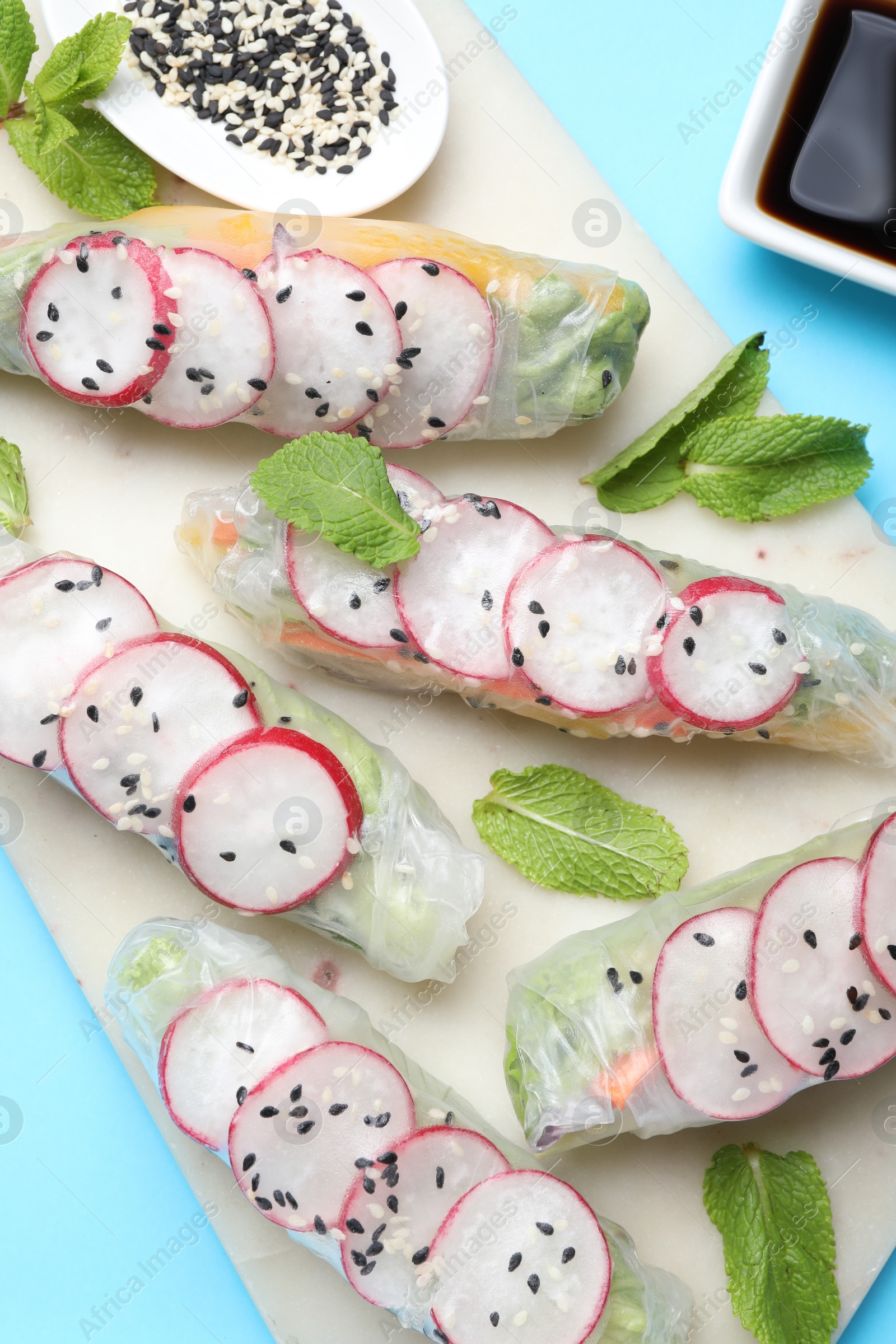 Photo of Delicious spring rolls, mint, sesame and soy sauce on light blue background, top view