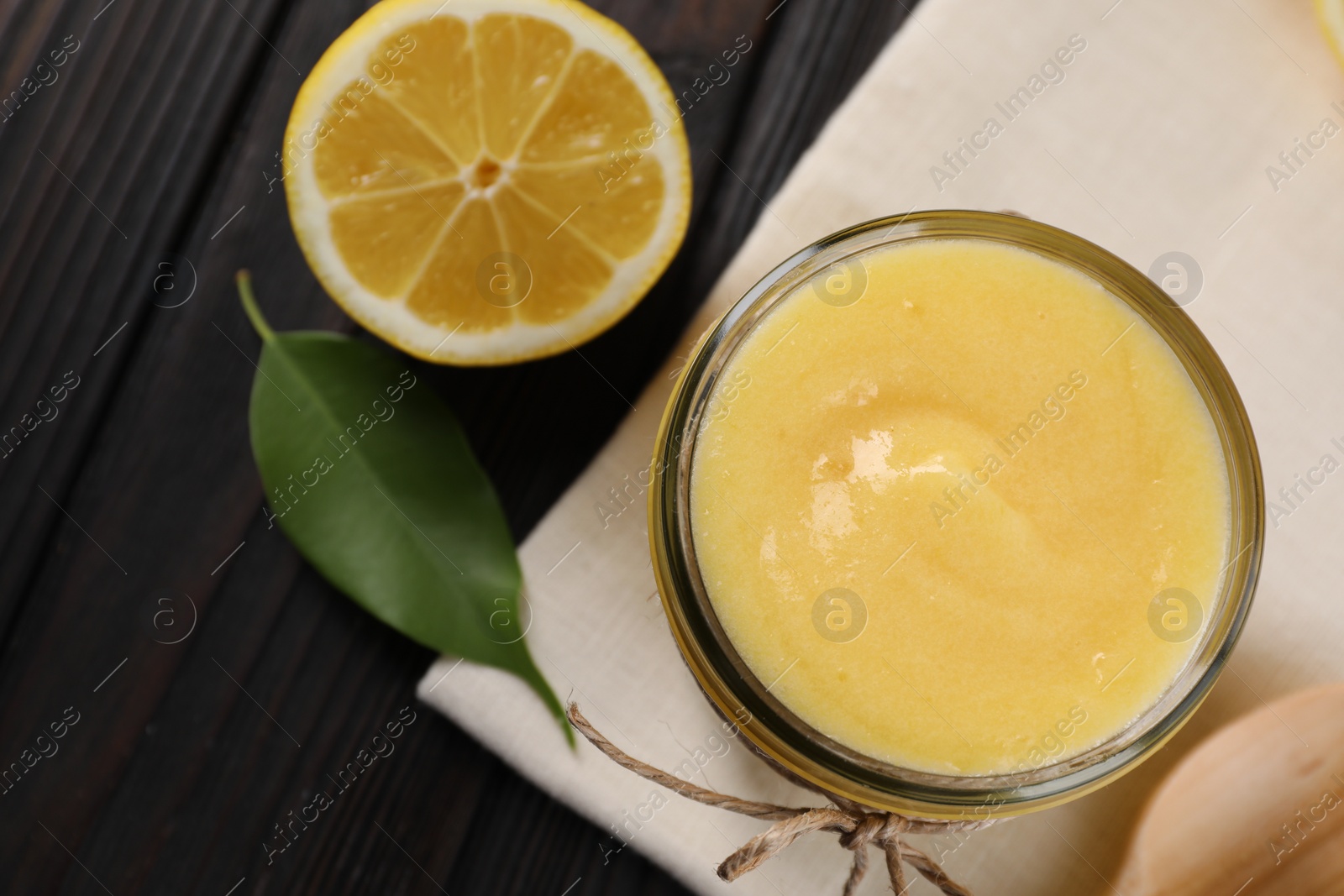 Photo of Delicious lemon curd in glass jar, fresh citrus fruit, green leaf and spoon on wooden table, top view