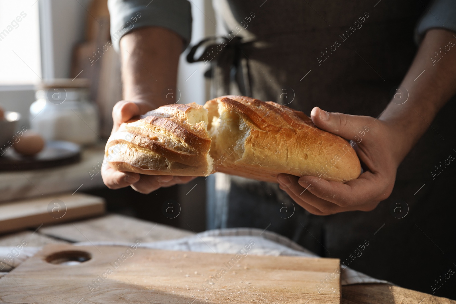 Photo of Man breaking loaf of fresh bread at wooden table indoors, closeup