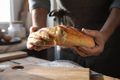 Photo of Man breaking loaf of fresh bread at wooden table indoors, closeup