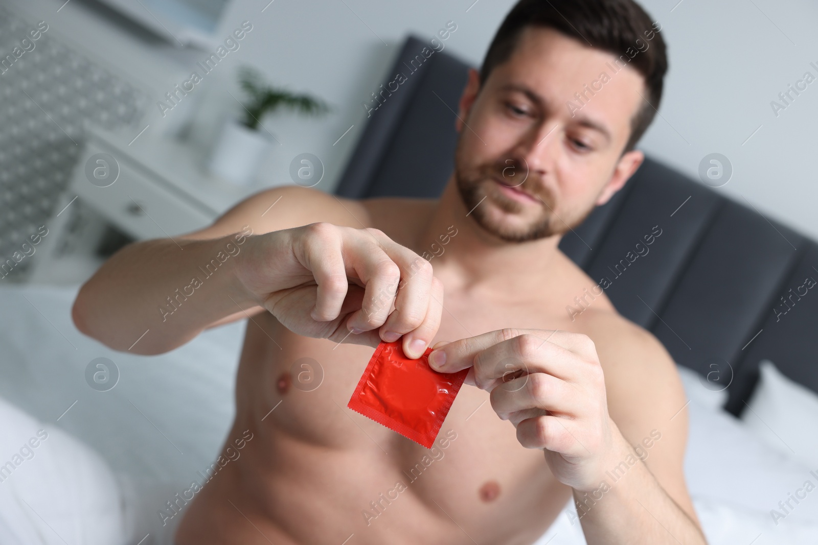 Photo of Man opening pack of condom on bed indoors, selective focus