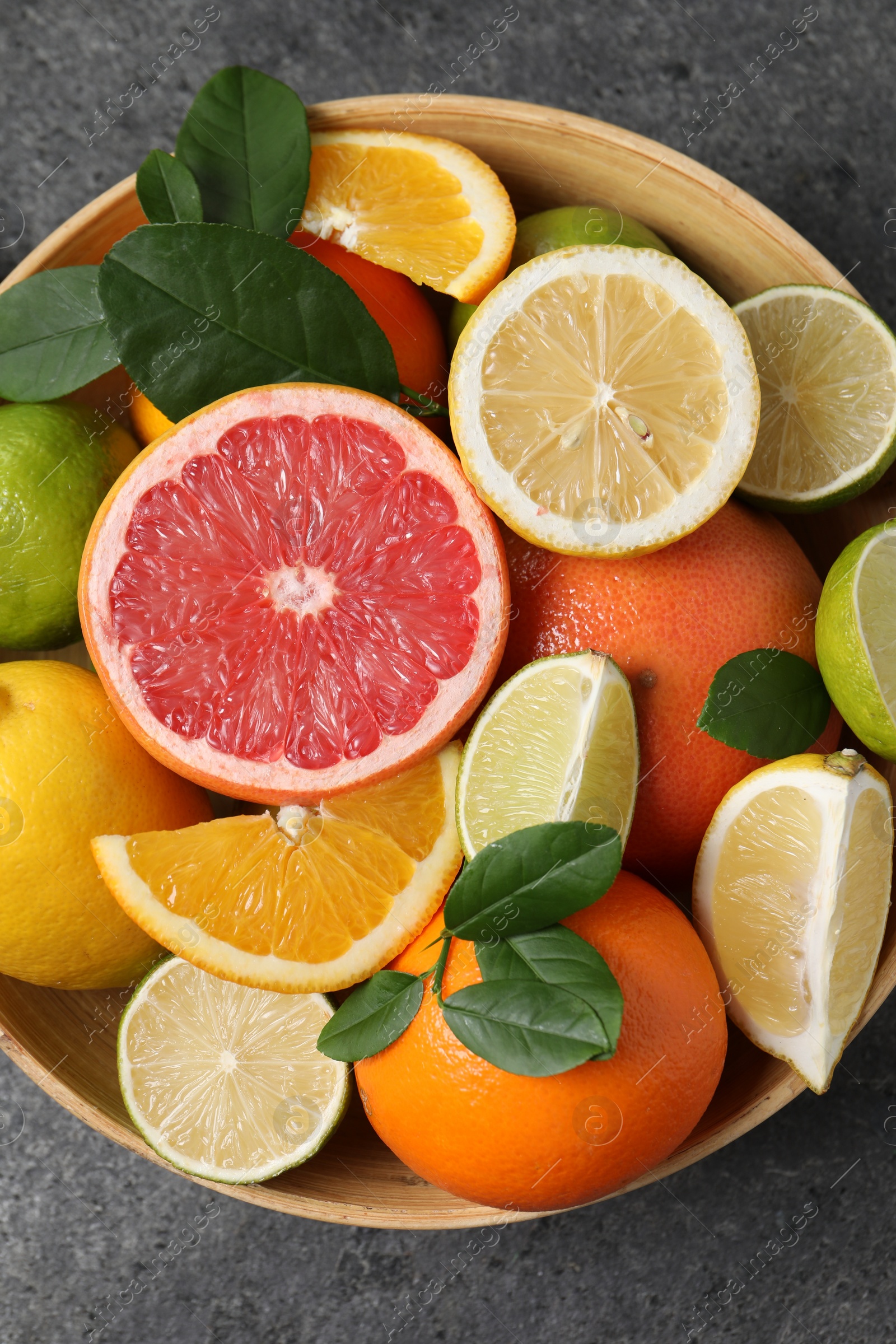 Photo of Different fresh citrus fruits and leaves in bowl on grey textured table, top view