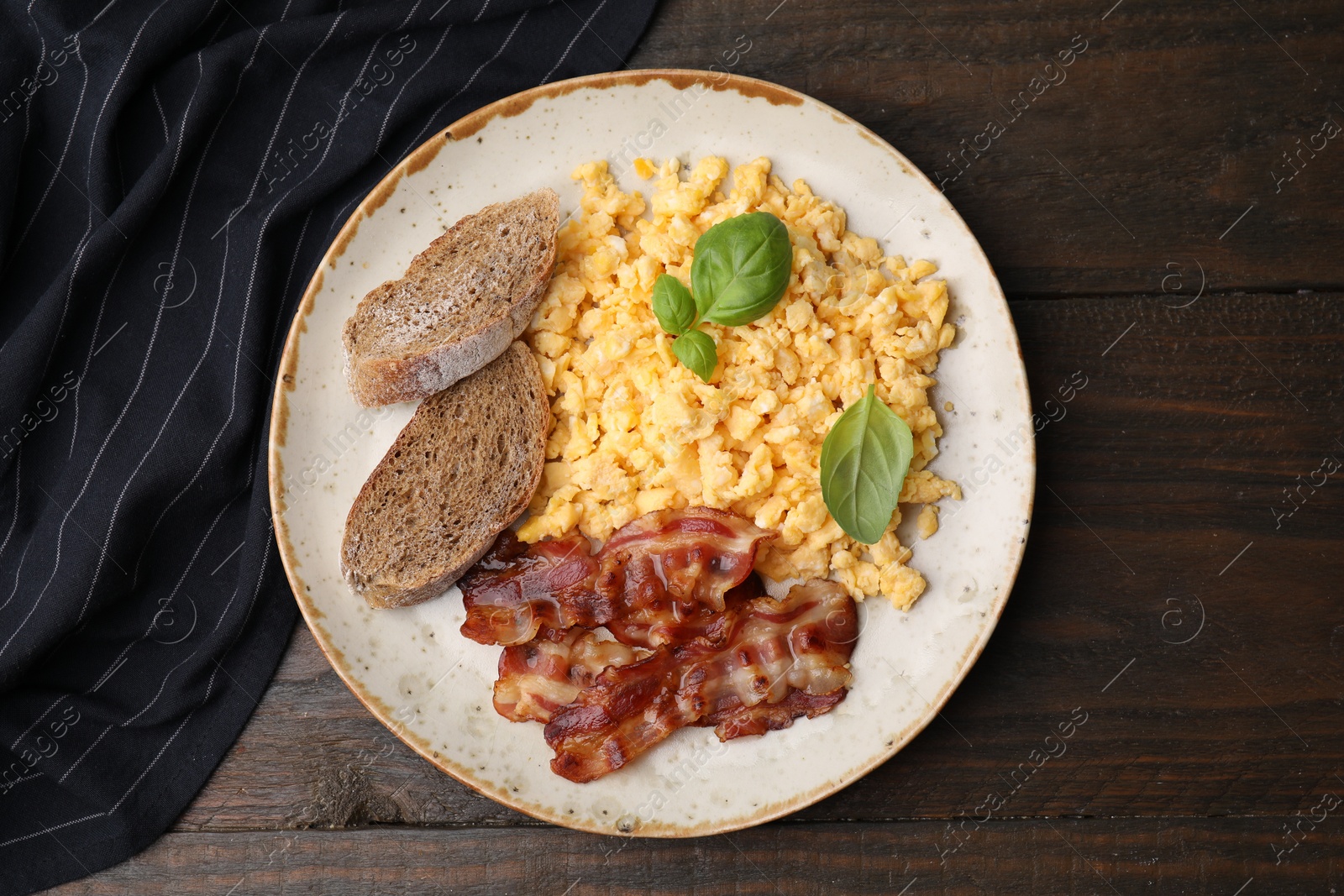 Photo of Delicious scrambled eggs with bacon and basil in plate on wooden table, top view