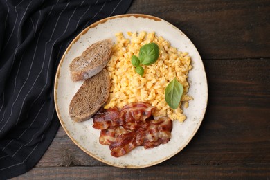 Delicious scrambled eggs with bacon and basil in plate on wooden table, top view