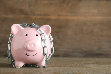 Photo of Piggy bank with steel chain on wooden table, space for text. Money safety concept