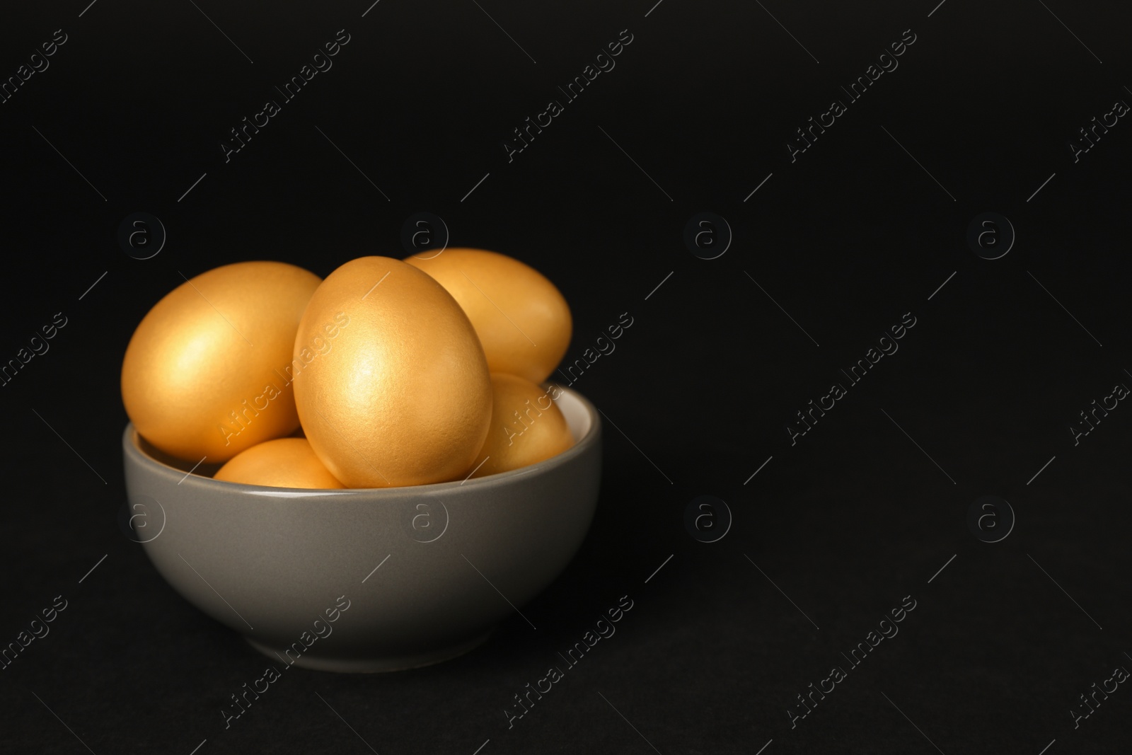 Photo of Shiny golden eggs in bowl on black background, space for text