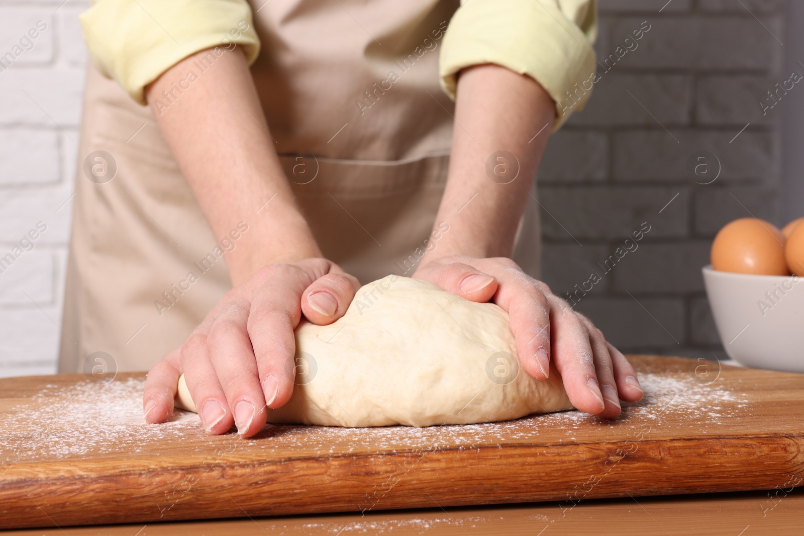 Photo of Woman kneading dough at wooden table near white brick wall, closeup