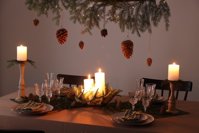 Photo of Christmas celebration. Cones hanging from fir tree branch over table with burning candles and tableware