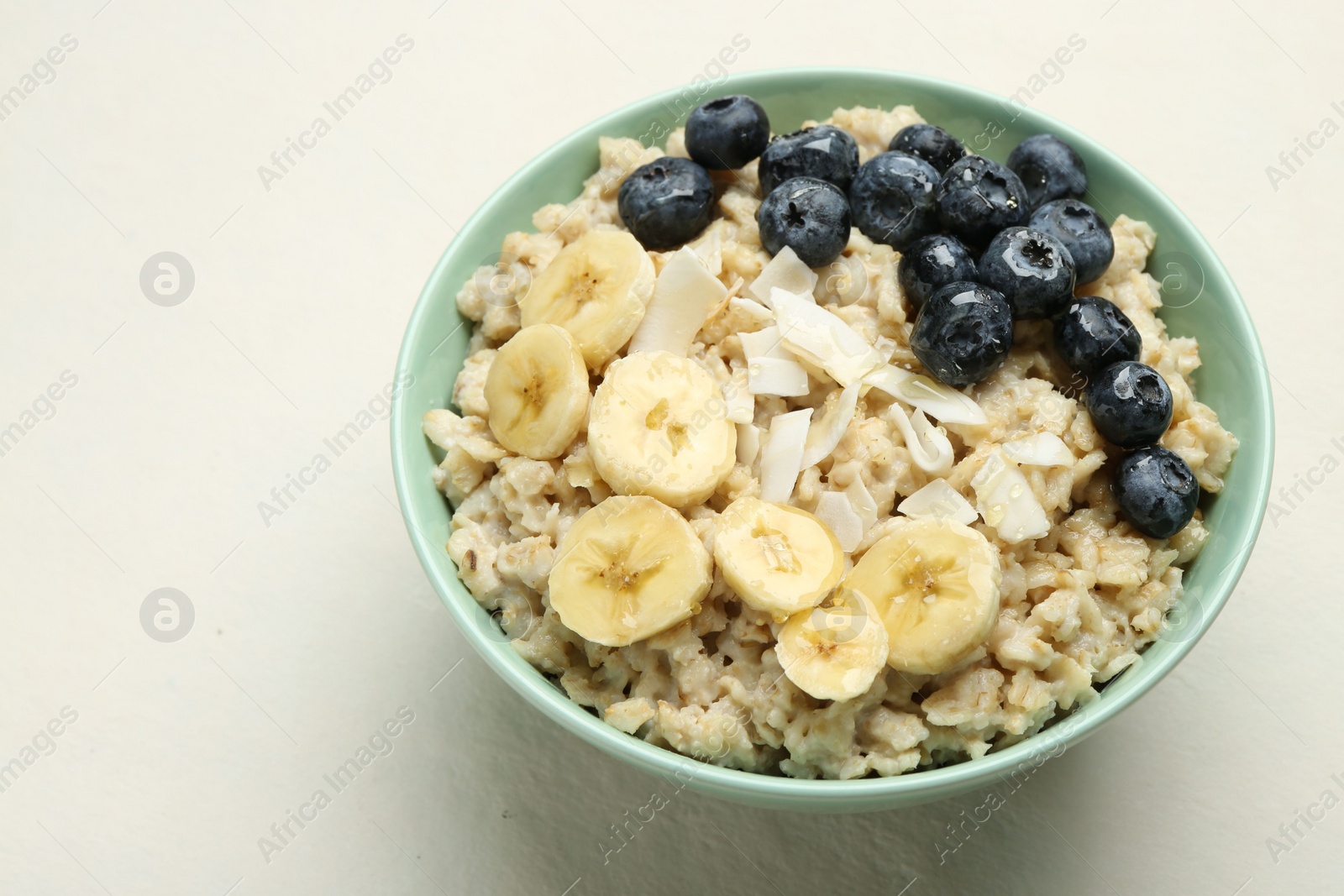 Photo of Tasty oatmeal with banana, blueberries, coconut flakes and honey served in bowl on beige table, space for text