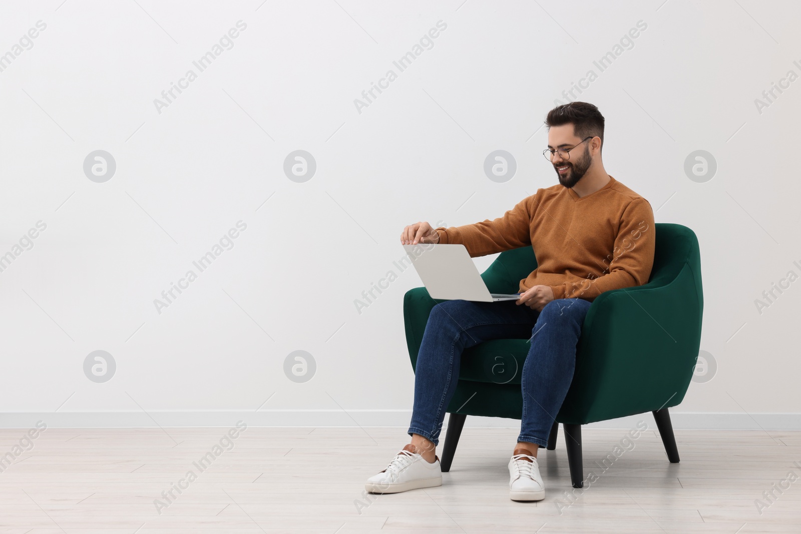 Photo of Handsome man using laptop while sitting in armchair near white wall indoors, space for text