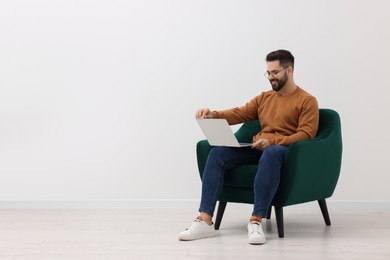 Handsome man using laptop while sitting in armchair near white wall indoors, space for text