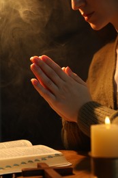 Woman praying at table with burning candle and Bible, closeup