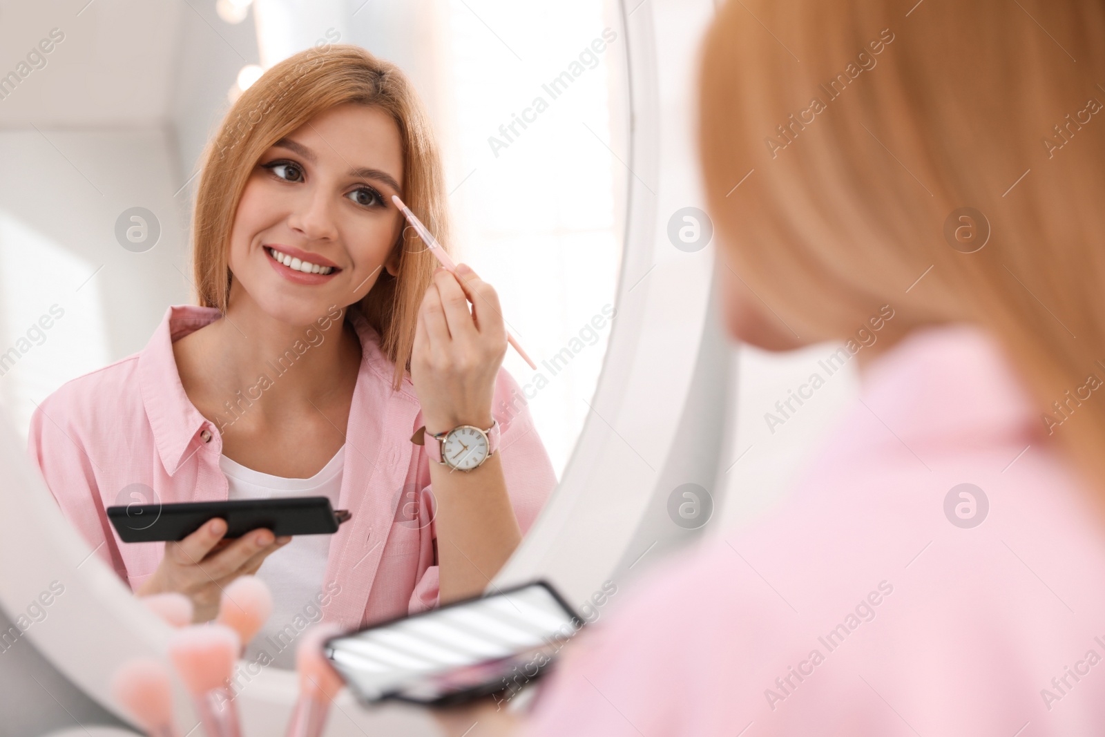 Photo of Beautiful young woman applying makeup near mirror indoors