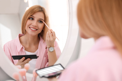 Beautiful young woman applying makeup near mirror indoors