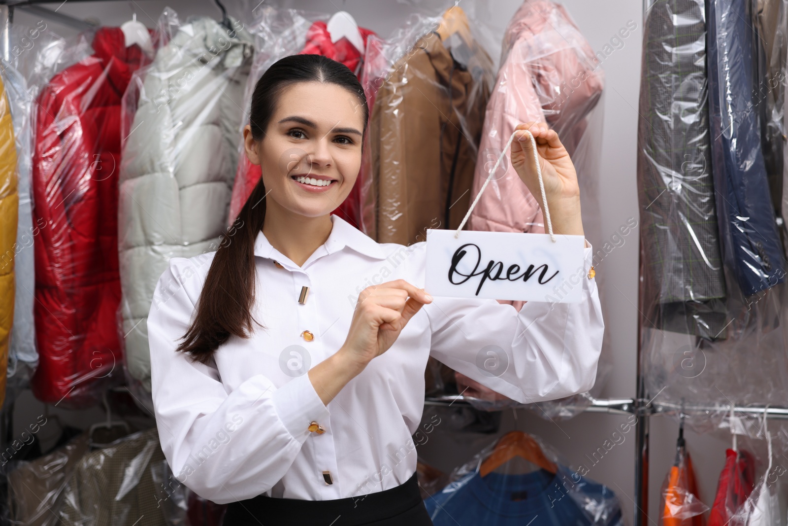 Photo of Dry-cleaning service. Happy worker holding Open sign near rack with clothes indoors