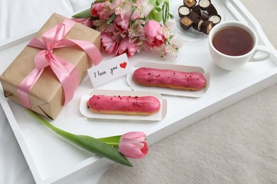 Photo of Tasty breakfast served in bed. Delicious desserts, tea, flowers, gift box and card with phrase I Love You on tray