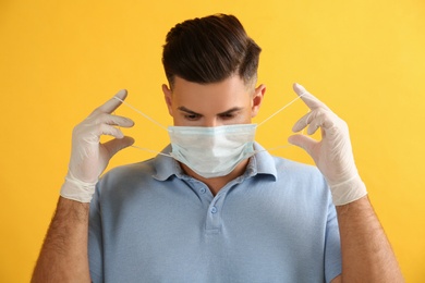 Man in medical gloves putting on protective face mask against yellow background