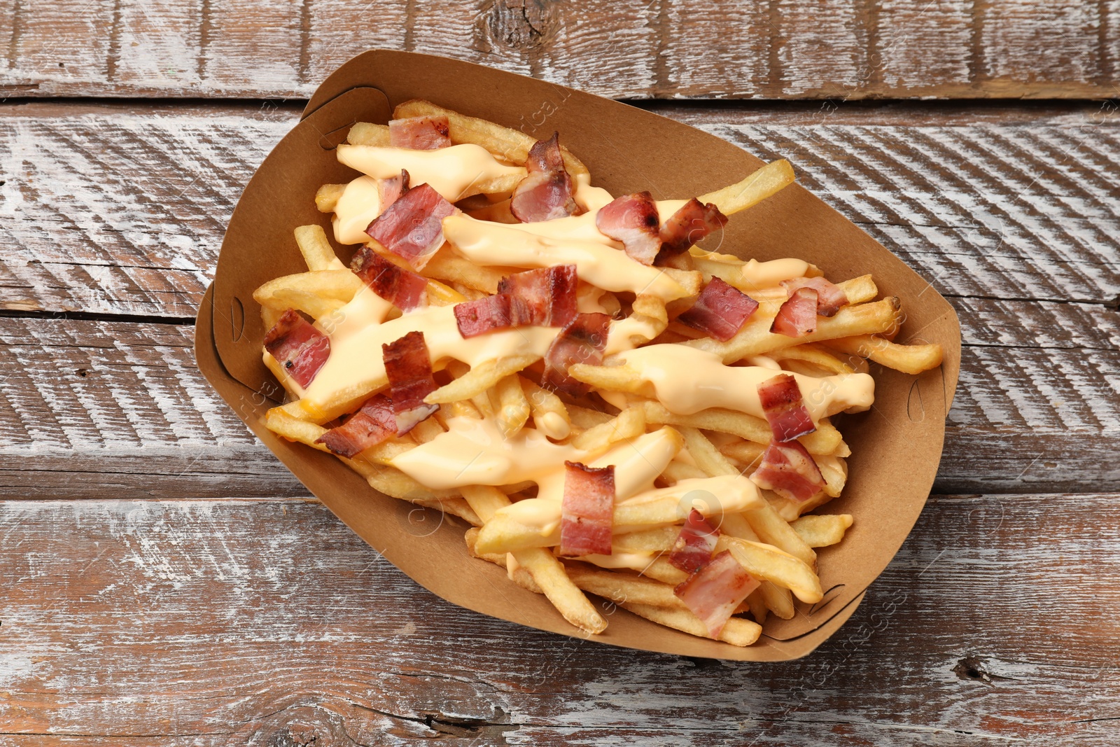 Photo of Tasty potato fries, cheese sauce and bacon in paper container on wooden table, top view