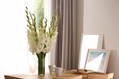 Vase with beautiful white gladiolus flowers, pictures and cups on wooden table in room, space for text