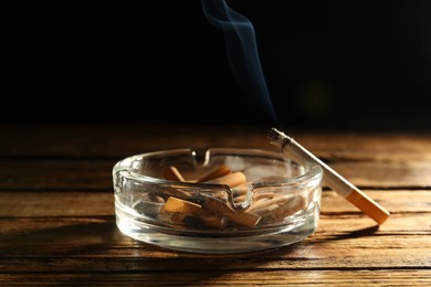 Photo of Glass ashtray with stubs and smoldering cigarette on wooden table against black background