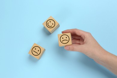 Image of Complaint. Woman choosing wooden cube with sad emoticon instead of other ones with drawn happy and neutral faces on light blue background, top view