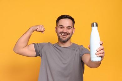 Photo of Happy man holding thermo bottle and showing arm on orange background