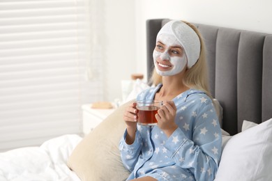 Young woman with face mask drinking tea on bed at home, space for text. Spa treatments