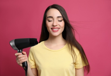 Beautiful young woman using hair dryer on pink background