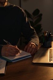 Man taking notes at wooden table indoors in evening, closeup