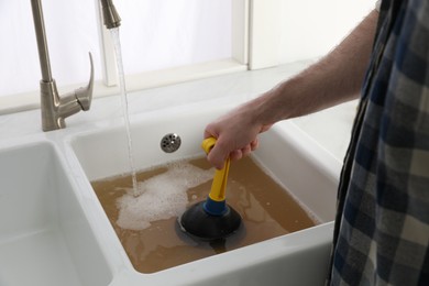 Photo of Man using plunger to unclog sink drain in kitchen, closeup