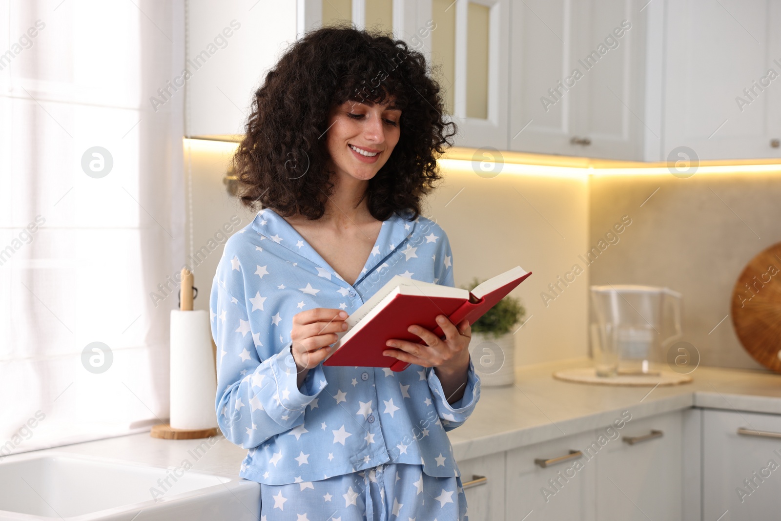 Photo of Beautiful young woman in stylish pyjama reading book in kitchen