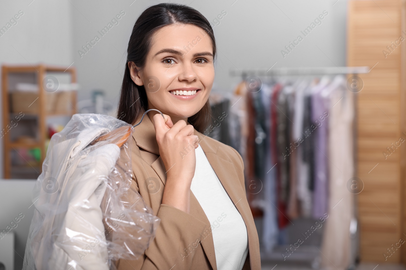 Photo of Dry-cleaning service. Happy woman holding hanger with jacket in plastic bag indoors, space for text