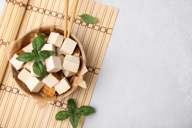 Photo of Bamboo mat with bowl of smoked tofu cubes and basil on white table, flat lay. Space for text